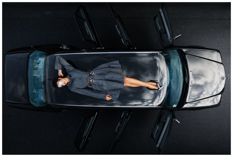 Aerial view of a woman lying on the transparent roof of a black car with open doors on an asphalt surface.