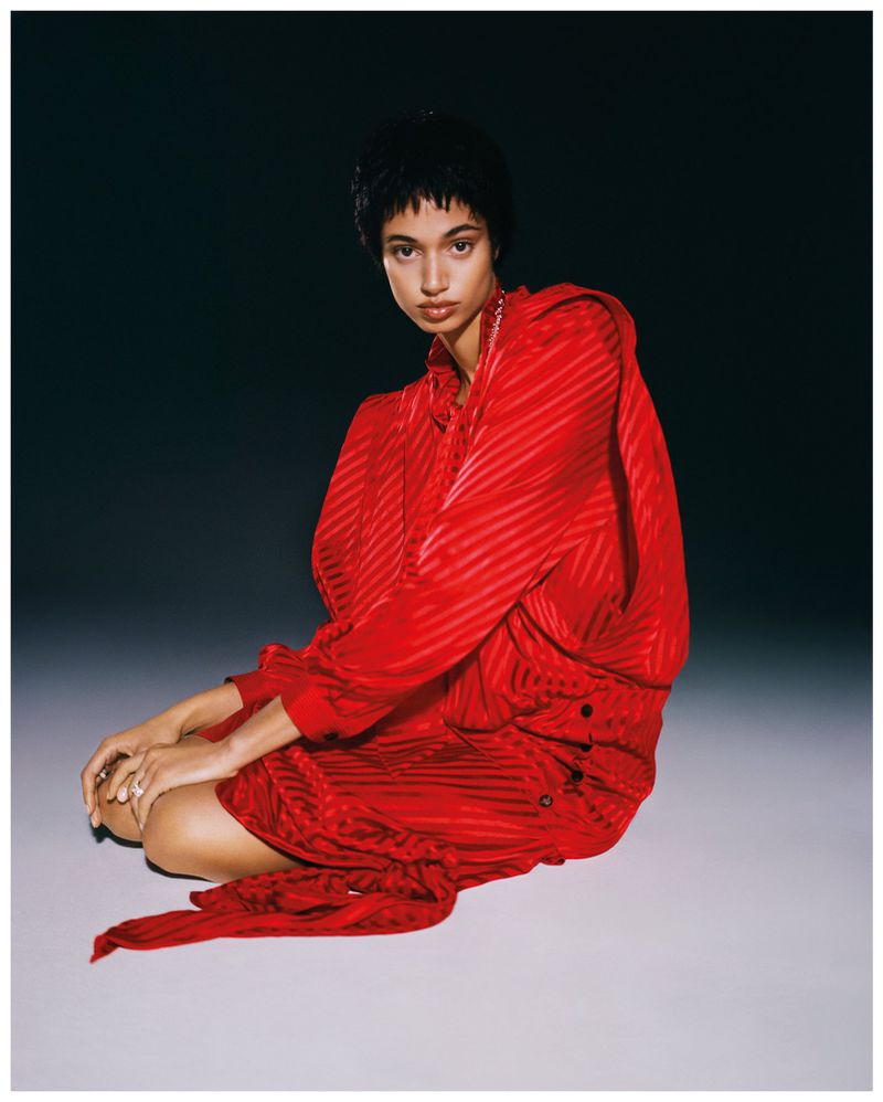 Woman in red striped dress sitting in photo studio, lit by studio lighting, posing for fashion photography. Dark backdrop, medium shot.