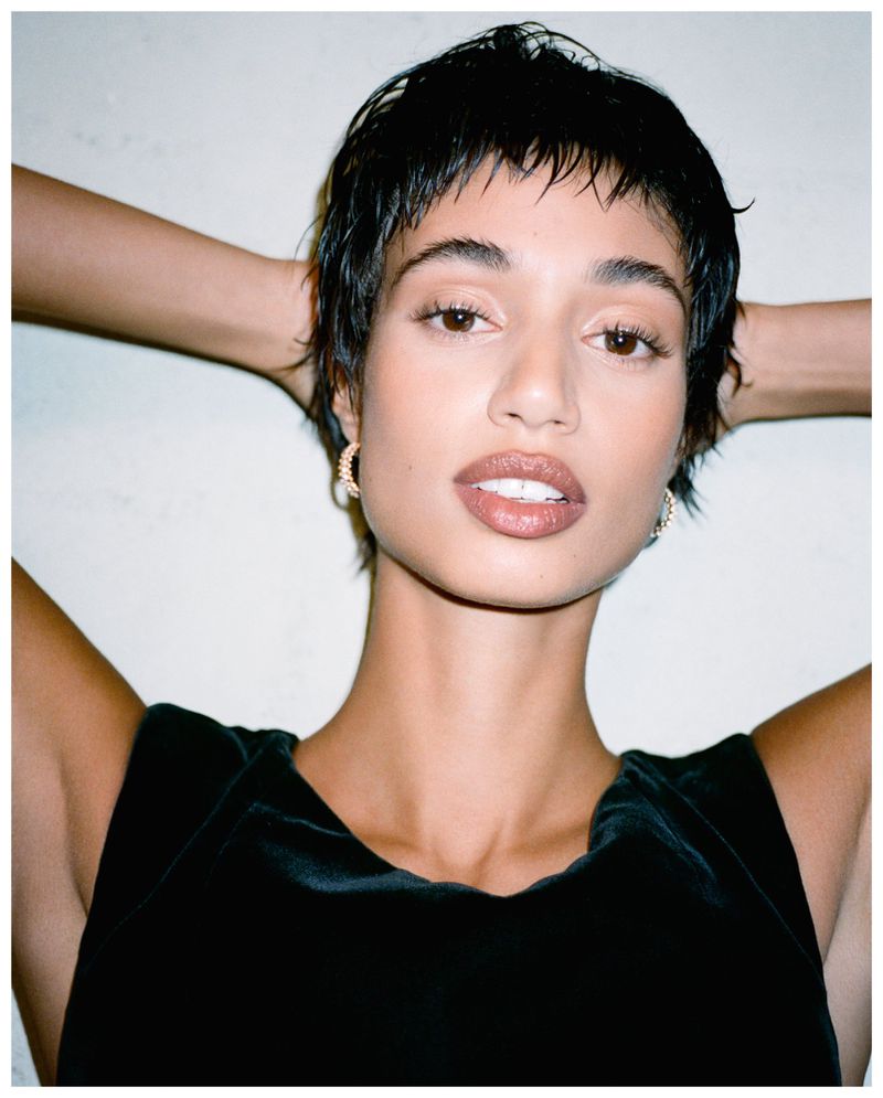 Close-up portrait of a young adult woman with dark hair, wearing a black top and hoop earrings, posing against a white background.