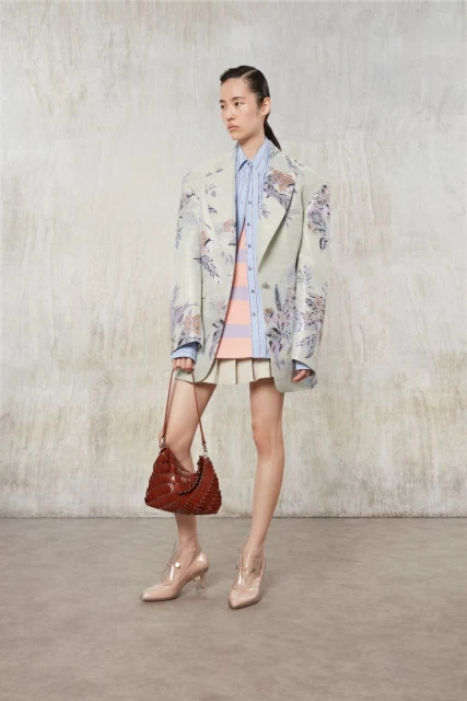 Elegant Asian woman in a patterned blazer and skirt, posing indoors with a brown purse and nude heels against a textured wall.