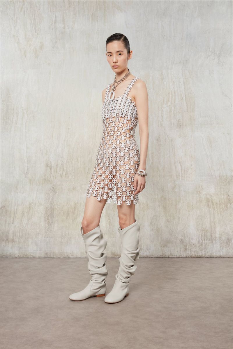 Elegant Asian woman poses in a sheer patterned mini dress, complemented by statement jewelry and slouchy boots against a textured wall.