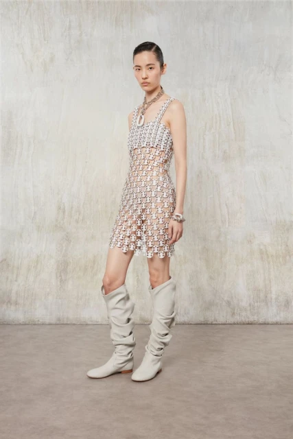 Elegant Asian woman poses in a sheer patterned mini dress, complemented by statement jewelry and slouchy boots against a textured wall.