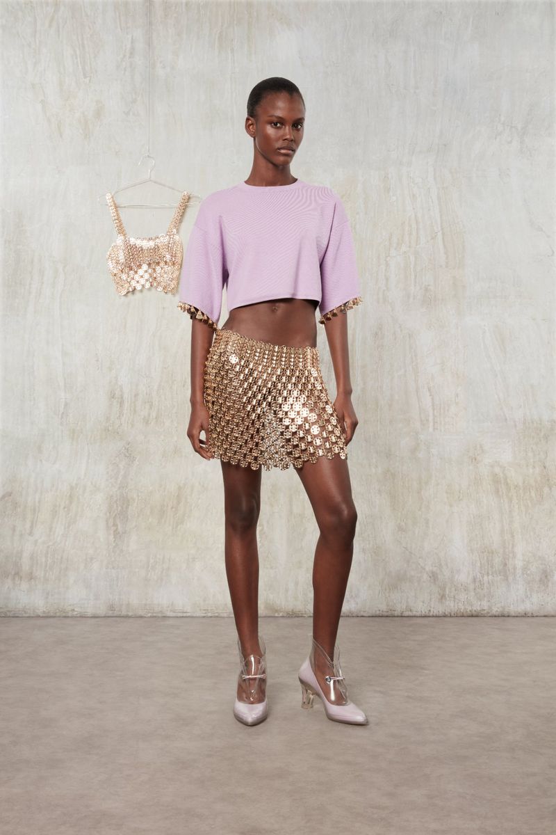 Elegant Black female model poses in a lilac top and gold chain skirt, complemented by a matching top and heels against a neutral backdrop.