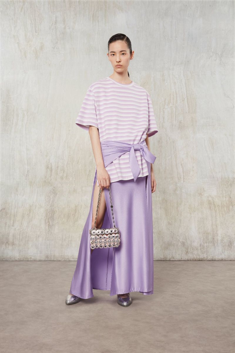 Indoor fashion shot of a young woman in a lilac striped shirt, lilac wrap, and long lilac skirt, holding a beaded purse.