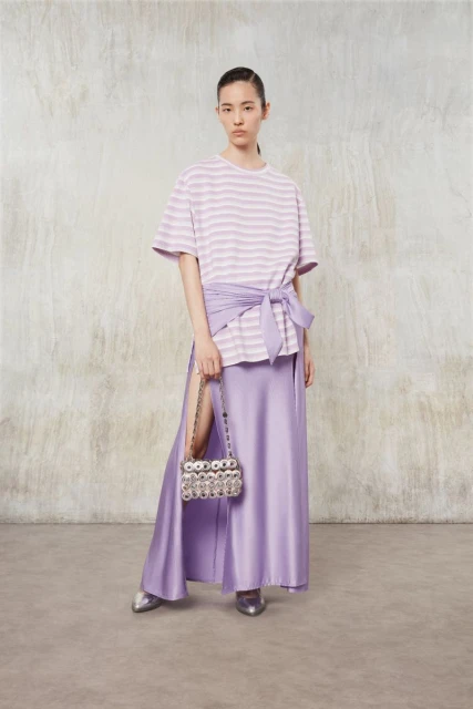 Indoor fashion shot of a young woman in a lilac striped shirt, lilac wrap, and long lilac skirt, holding a beaded purse.
