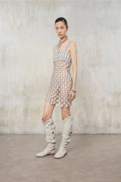 Model wearing a metallic chain-link dress and slouchy light boots against a textured concrete wall