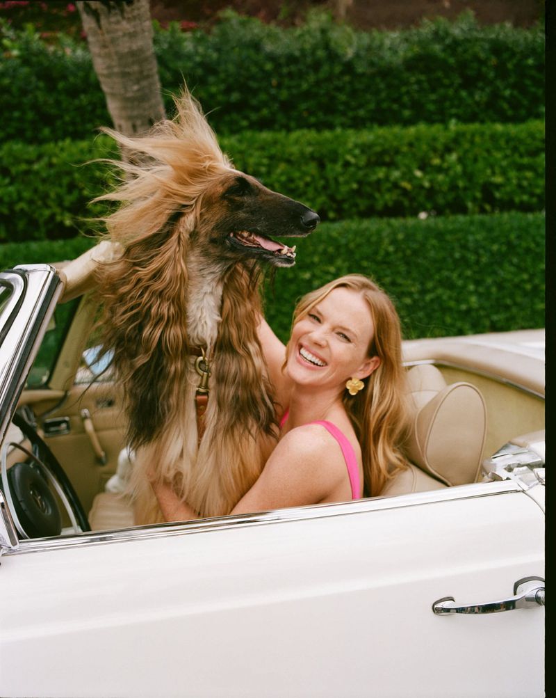 Smiling woman in pink with Afghan Hound dog in white convertible car against green hedge. Friendship, happy mood, and luxury lifestyle.