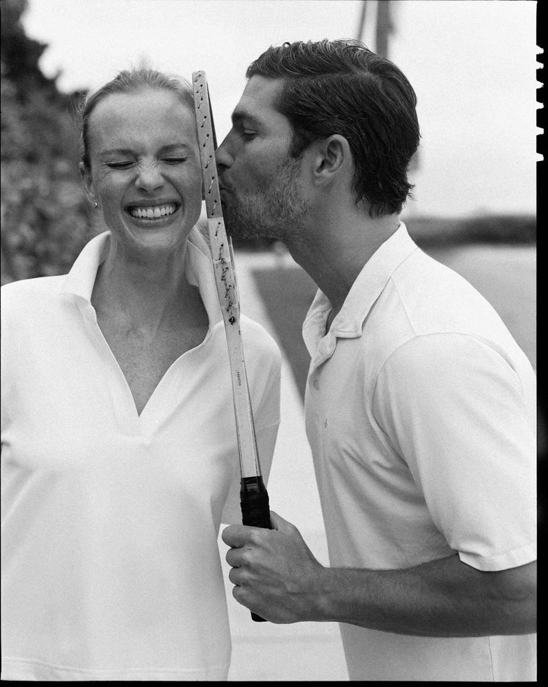 Monochrome photo of a smiling woman holding a tennis racket as a man kisses it near her face, both in polo shirts outdoors.