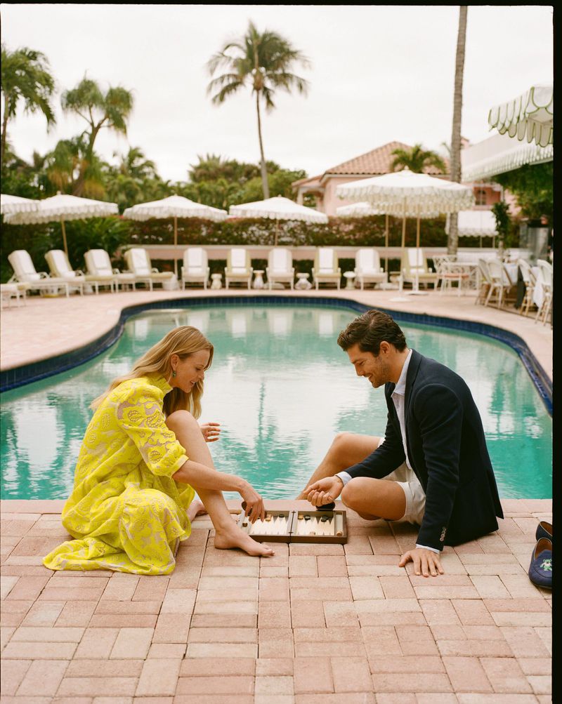 A man and woman playing backgammon poolside at a luxury resort. Palm trees, chairs with umbrellas, and a building are visible.