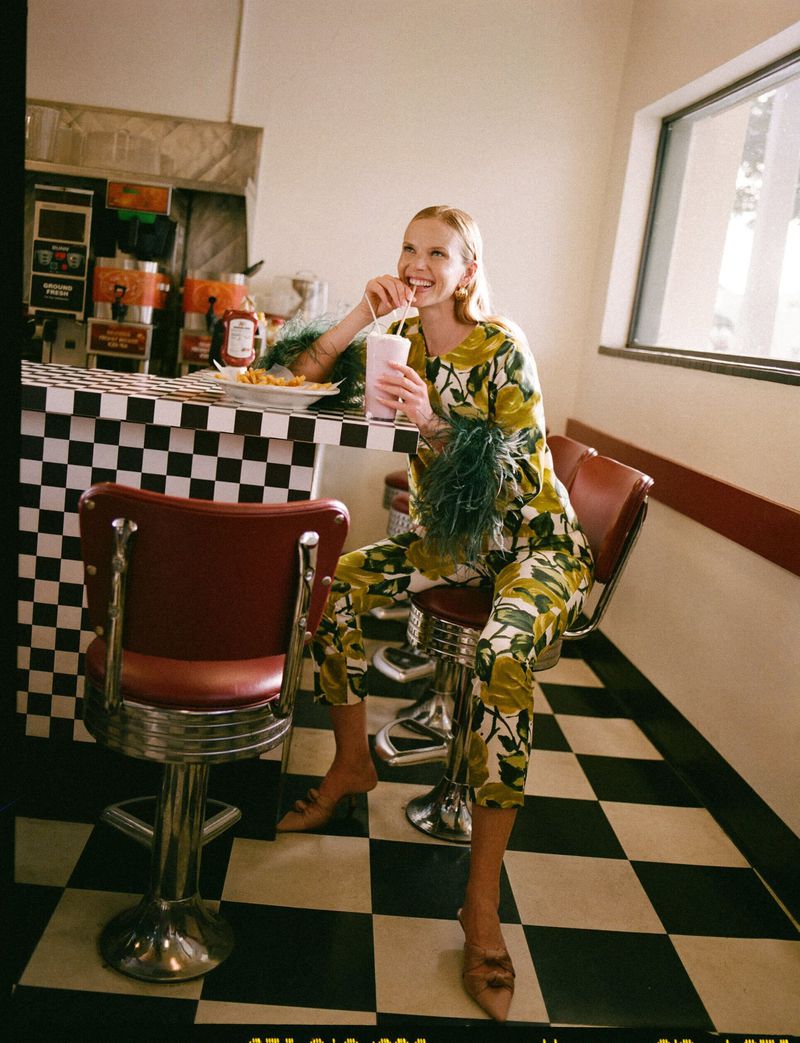 A woman in a floral outfit enjoys a milkshake and fries at an old-fashioned diner with checkered floors and red swivel chairs.