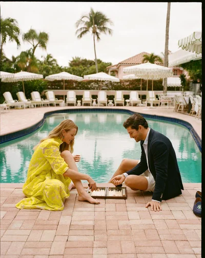 Anne Vyalitsyna in a yellow dress and Benj Lee in a navy blazer playing backgammon by a resort swimming pool.