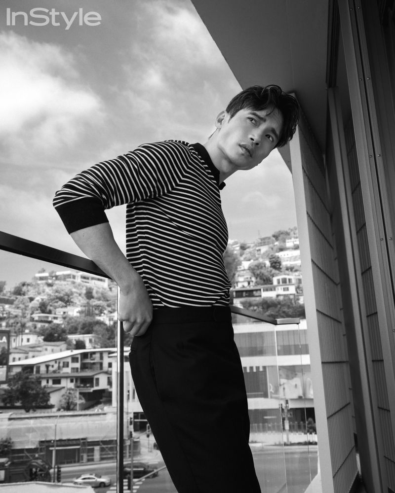 Black and white fashion editorial shot of an Asian male model in a striped shirt leaning against a balcony overlooking hillside homes.