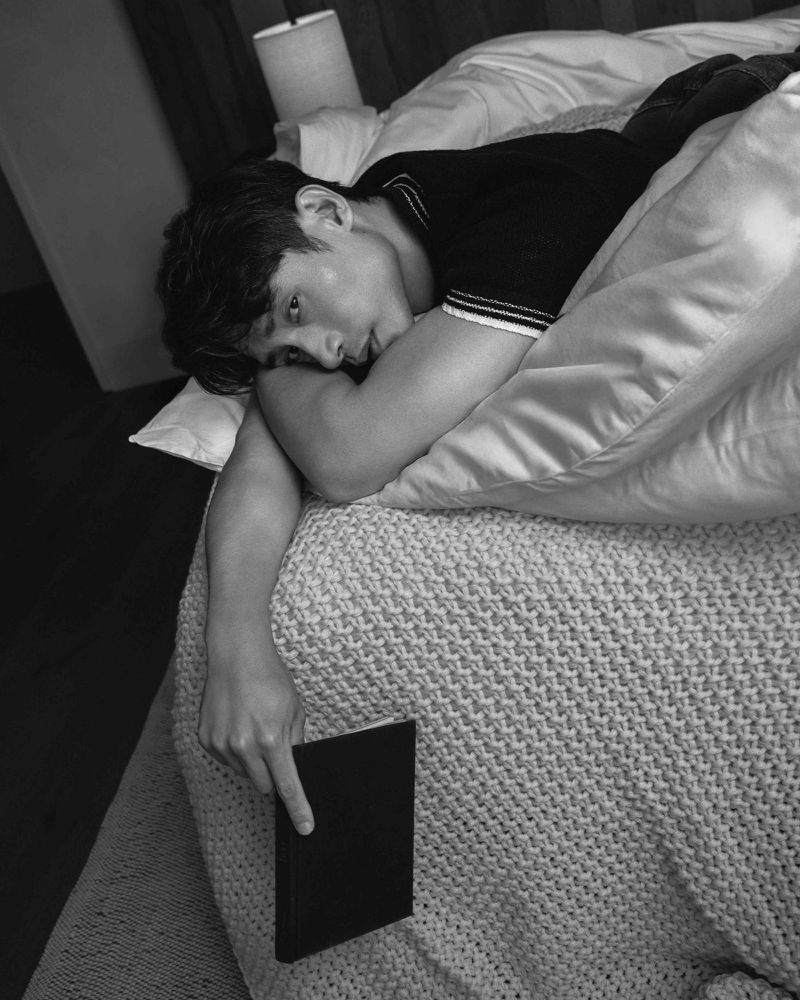 Monochrome portrait of a young man lying in bed, resting on his arms and holding a closed book, with a lamp visible in the background.