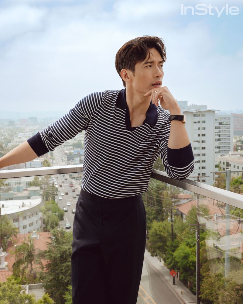 Adult man in striped shirt poses on balcony in an urban setting with city buildings and trees. Fashion editorial shot with natural light.