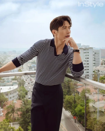 Adult man in striped shirt poses on balcony in an urban setting with city buildings and trees. Fashion editorial shot with natural light.