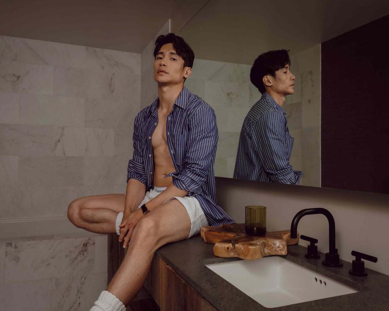Attractive Asian man poses in bathroom wearing blue striped shirt and white shorts, reflected in mirror behind a dark countertop.
