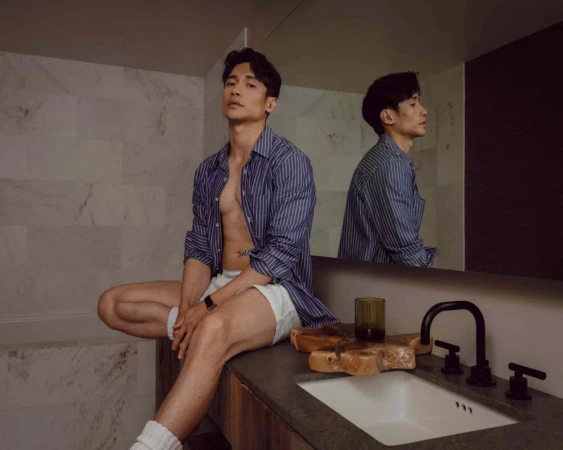 Attractive Asian man poses in bathroom wearing blue striped shirt and white shorts, reflected in mirror behind a dark countertop.