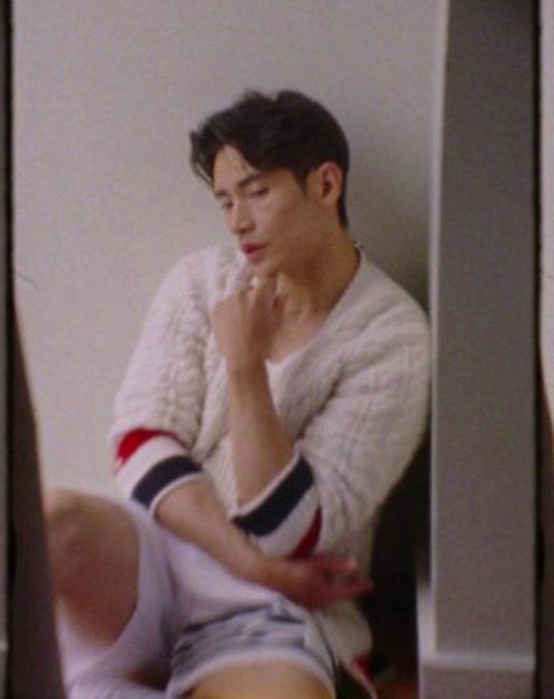 Young man sitting indoors wearing a white sweater with blue and red stripes on the sleeves and grey shorts, looking downwards. 