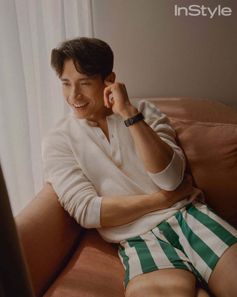 Smiling adult male poses indoors in a white shirt and striped shorts, resting his hand near his face, magazine logo visible.