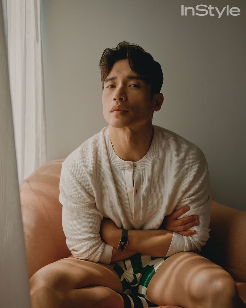 An adult male poses indoors, sitting on a pink chair with his arms crossed. He is wearing a light-colored shirt and patterned shorts.