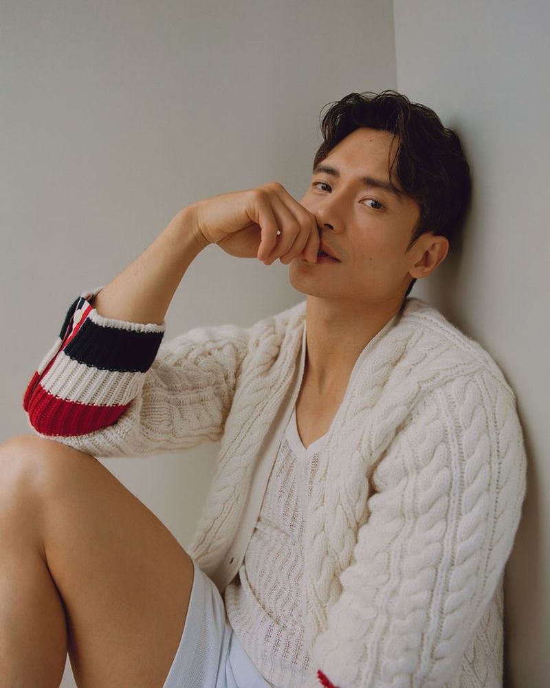 Man poses indoors wearing an open white cable knit cardigan and matching v-neck t-shirt with his hand touching his face.