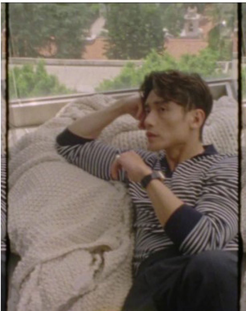 An adult male lounges on a couch with a knitted blanket near a window with greenery outside; he is wearing a striped top.