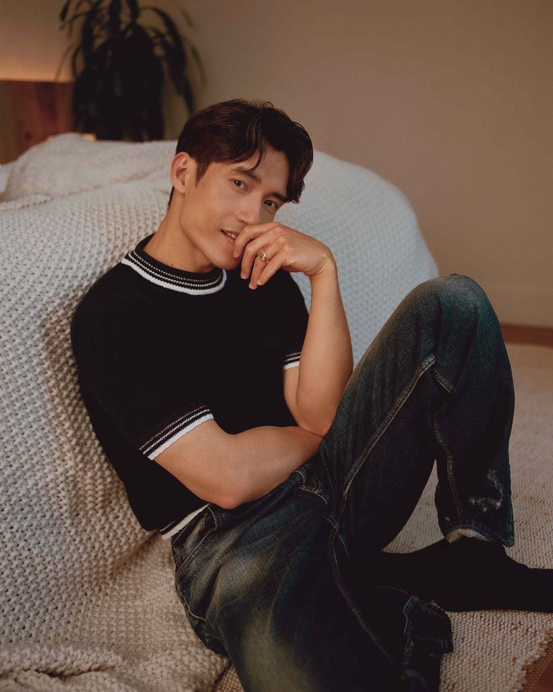 Attractive brunette man in dark jeans and short-sleeved polo shirt seated on a gray knitted bedspread and white wall.
