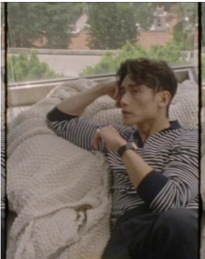 Manny Jacinto wearing a striped long-sleeve shirt reclining against a chunky knit blanket beside a large window.