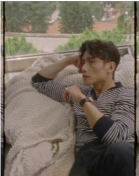 Manny Jacinto wearing a striped long-sleeve shirt reclining against a chunky knit blanket beside a large window.