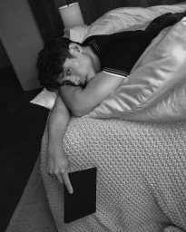 Manny Jacinto lying on a bed resting his head on his arm while holding a dark book in a black and white portrait.