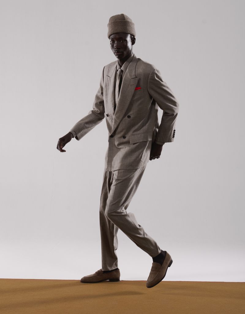 Stylish dark-skinned man in grey double-breasted suit and beanie confidently strides forward against a bright studio backdrop.