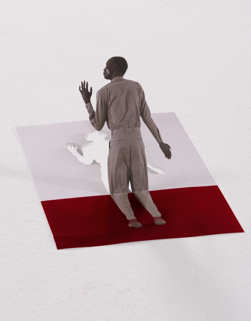 A man stands on a geometric red, white, and light-grey background, looking left with his arms outstretched on a white background.
