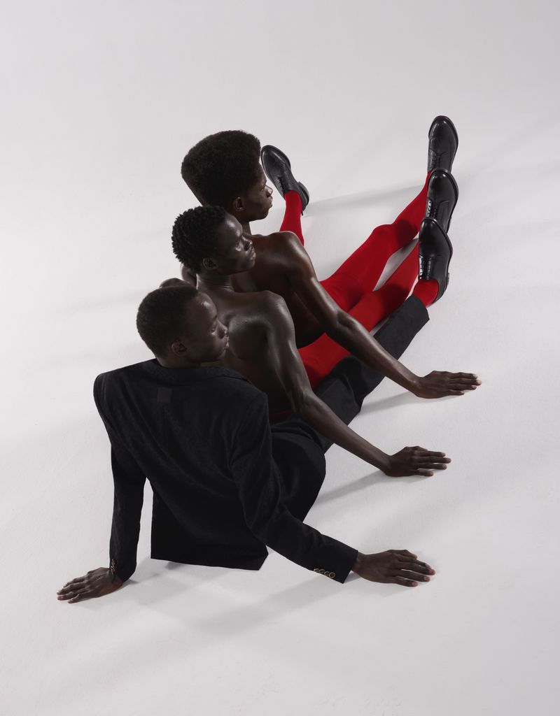 Three Black male fashion models in red tights, black pants, and a suit jacket pose in a line on a white backdrop in a studio shot.
