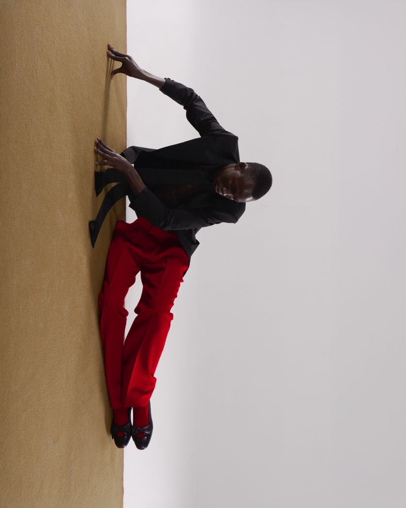 An African American male fashion model strikes a unique pose on the floor wearing a black jacket and red pants in a photo studio.
