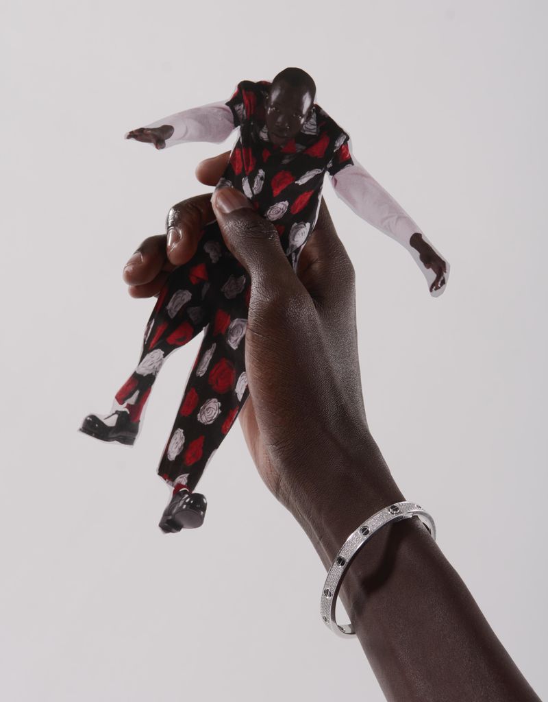 A dark-skinned hand holds a Black doll wearing a patterned suit against a white background, accessorized with a silver bracelet.