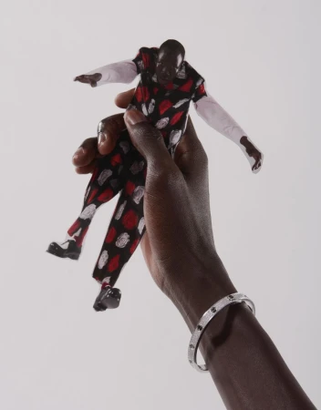A dark-skinned hand holds a Black doll wearing a patterned suit against a white background, accessorized with a silver bracelet.