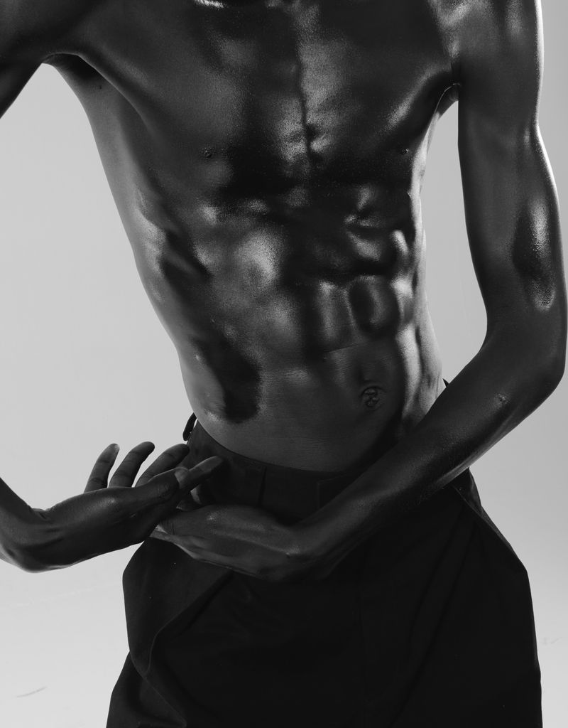 Dramatic close-up of a muscular African-American male torso with glossy black paint against a grey background in a studio setting.