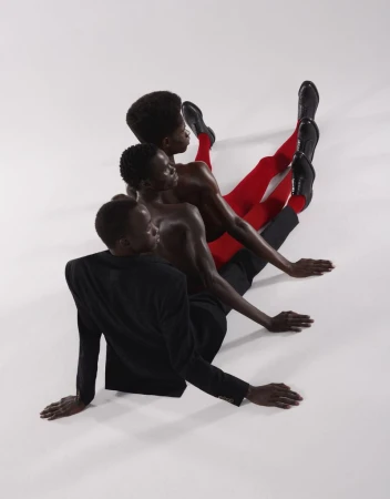Three Black male models seated diagonally on a white seamless backdrop wearing a black blazer, red tights, and dress shoes.