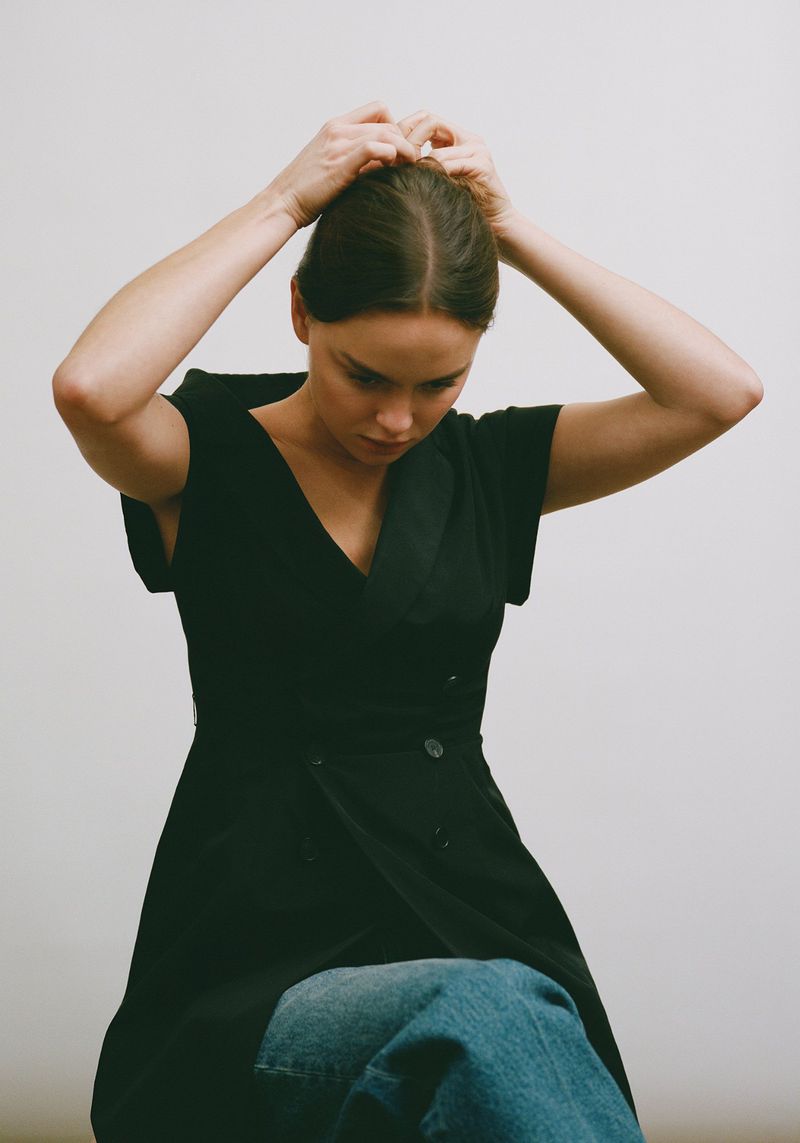 Indoor portrait shot of a woman with her hands on her head, wearing a black dress and jeans, against a plain white background.