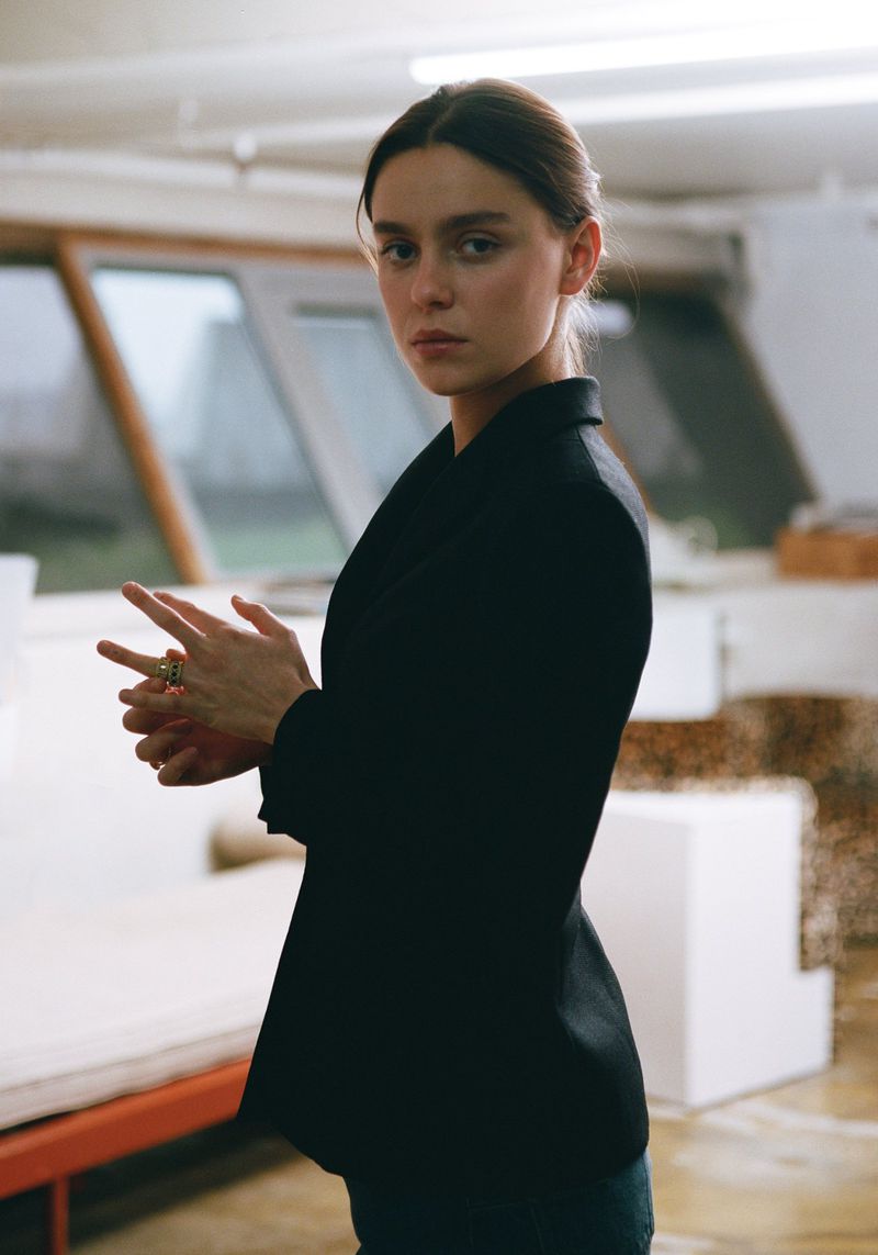 A young adult woman in a black blazer and jeans poses indoors with hands clasped, looking at the camera in a medium studio shot.