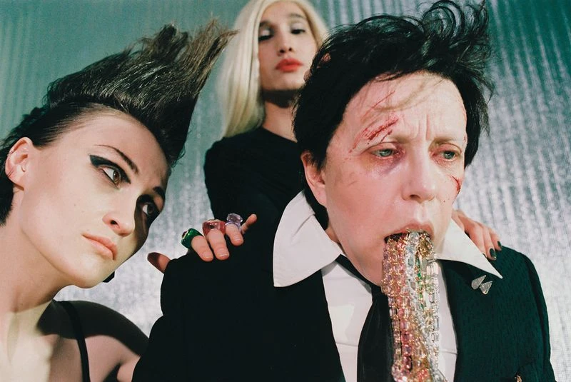 Unconventional studio portrait of three people: one with simulated cuts and jewels in their mouth, one with a bold haircut, and one with blonde hair.