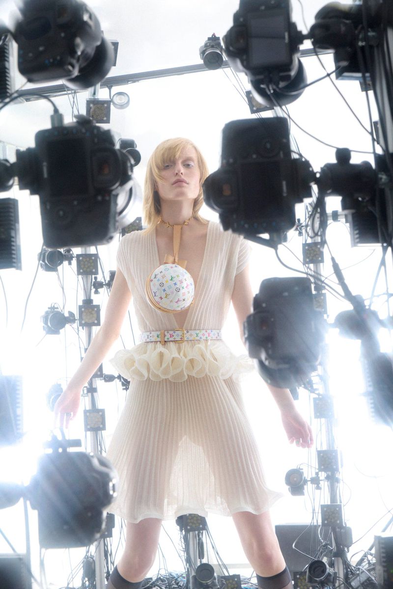 Fashionable adult female poses in a pleated dress and accessory, surrounded by many cameras under bright photo studio lights.