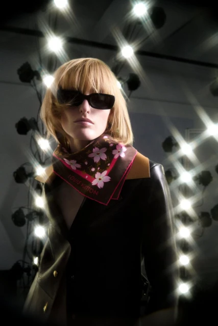 A woman wearing sunglasses and a patterned scarf is posing in front of a light array. The scene is lit dramatically with a spotlight effect.