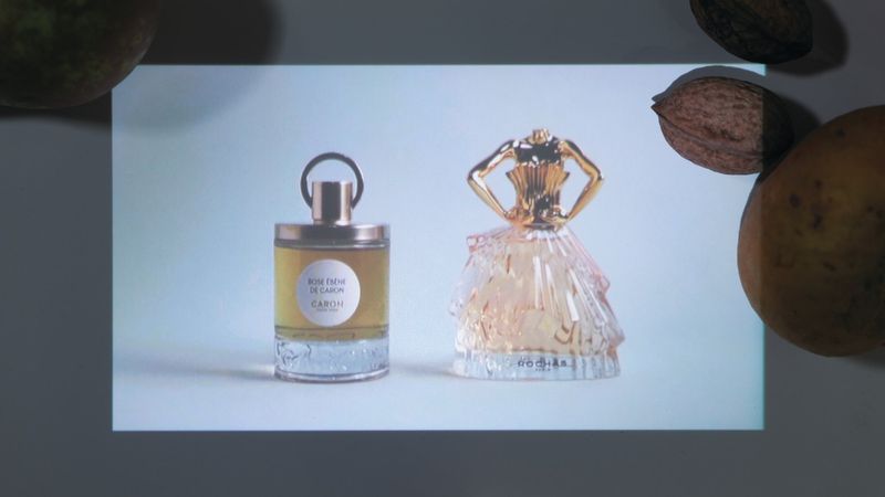 Still life of two perfume bottles, one shaped like a dress, on a white surface, surrounded by nuts and fruit.