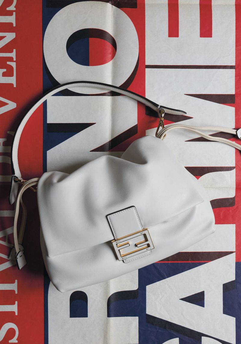 White leather shoulder bag with a gold buckle on a background with red, white, and blue abstract graphic shapes and patterns.