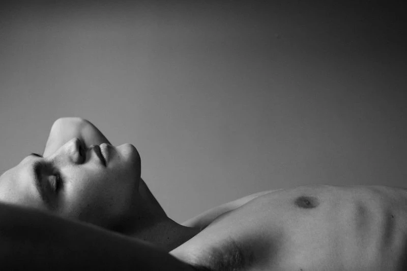 Monochrome portrait of a shirtless adult male lying down with his hand behind his head against a seamless grey background in a studio.