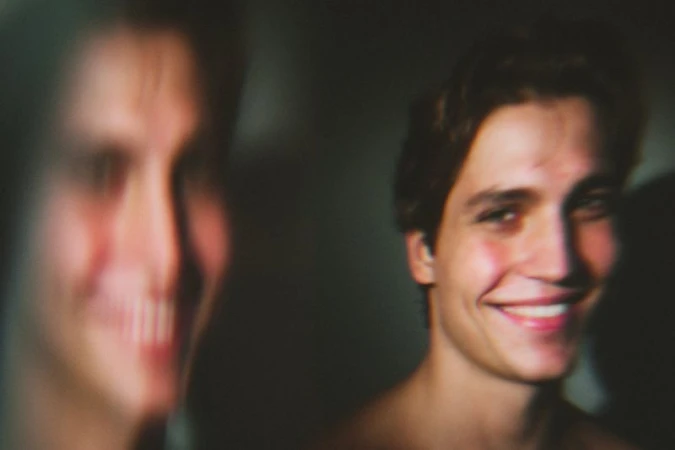 Close-up portrait of a smiling young man with bare shoulders next to a blurred smiling figure in dim lighting.