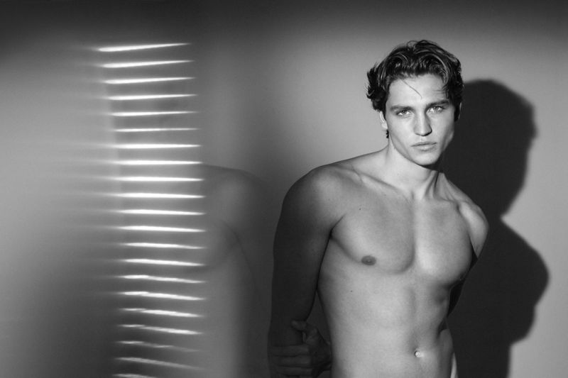 Black and white portrait of a shirtless adult male with medium-length wavy hair in front of a grey wall with window blind light patterns.