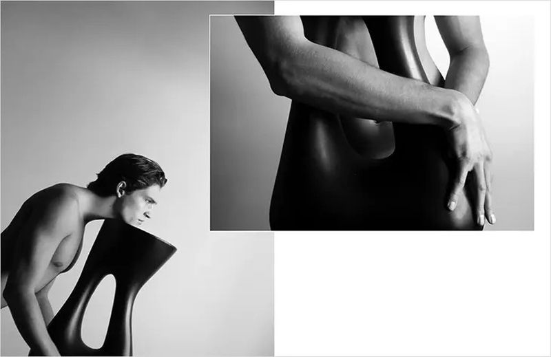 Black and white studio shot of a muscular adult male partially leaning on a sculptural black chair against a plain background.