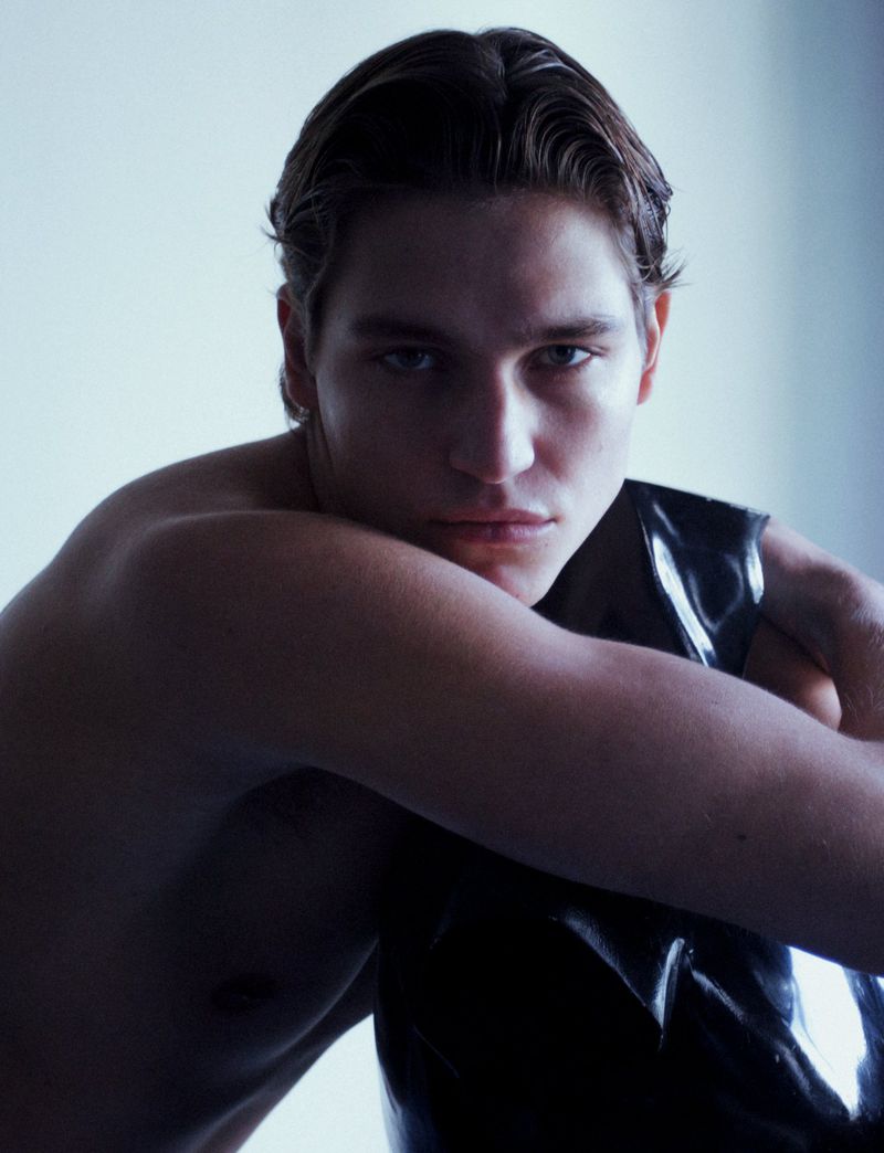 Close-up of a shirtless adult male with dark hair, posing indoors against a white background while wearing black shiny pants.